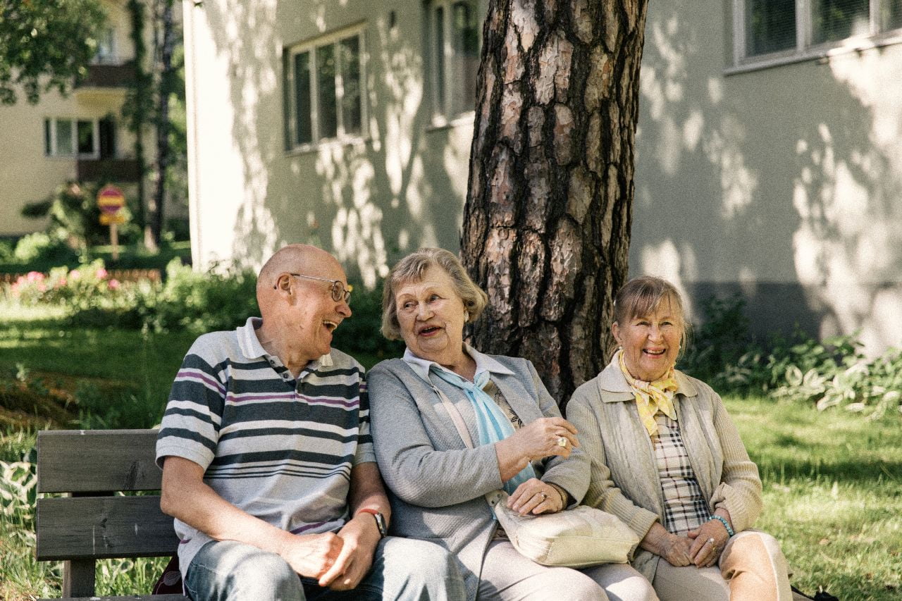 Kolme senioria nauraa yhdessä ulkona penkillä.