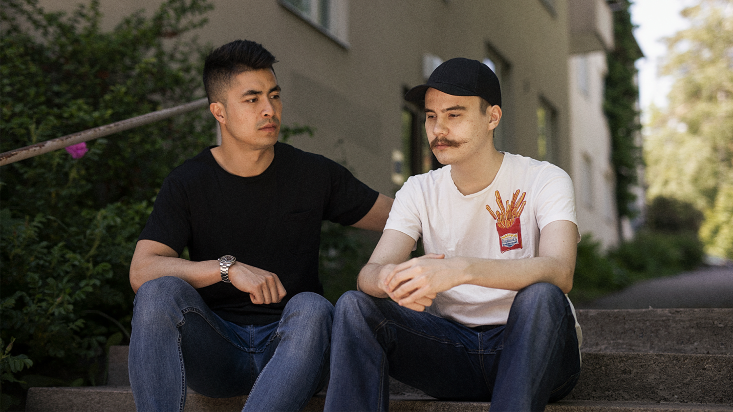 Kaksi nuorta istuu rappusilla. Toinen pitää kättä alakuloisen nuoren olalla lohduttavasti.