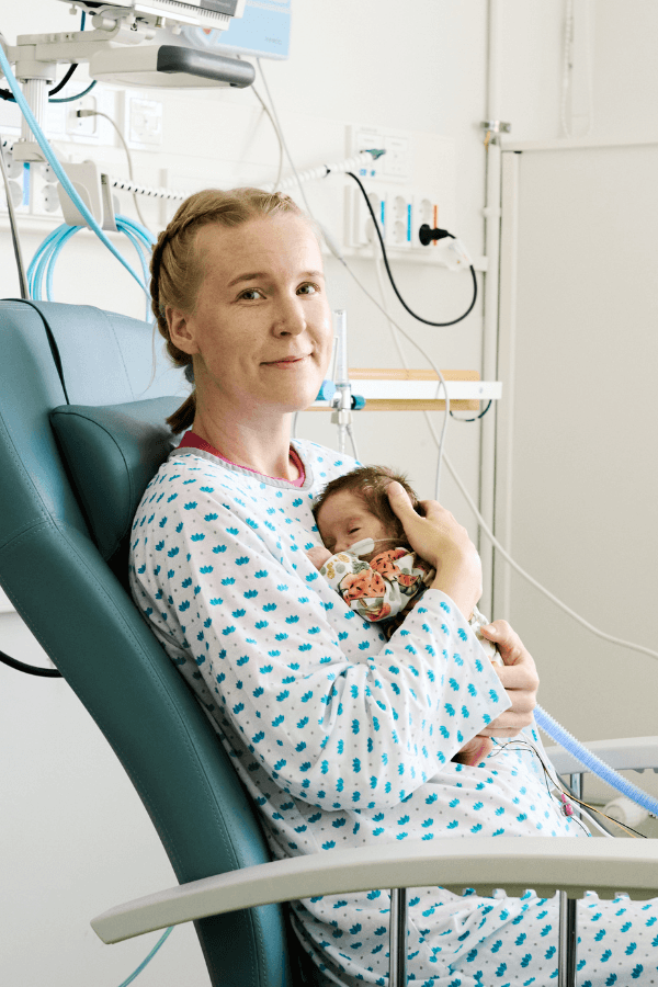 HelsinkiMission vapaaehtoinen perhekaveri hymyilee kameralle sylissään pieni vauva ja taustalla sairaalahuone.
