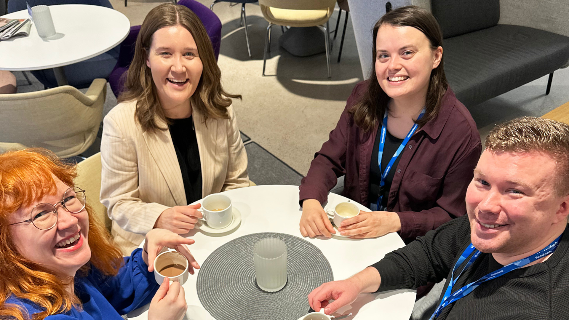 Neljä työntekijää istuu kahvitauolla yhteisen pöydän äärellä ja hymyilee iloisesti kameralle.