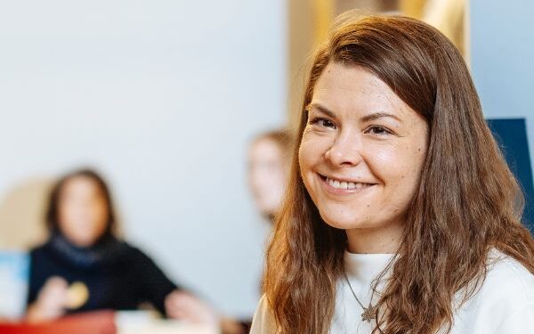 Nainen hymyilee ja katsoo kameraan kahvikuppi kädessään, taustalla kaksi ihmishahmoa istuu pöydän äärellä.