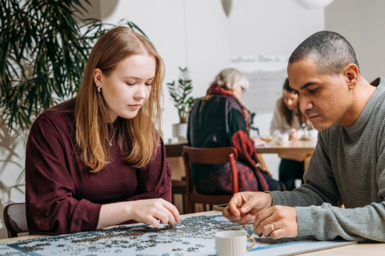 Kaksi ihmistä kokoaa palapeliä yhdessä ja taustalla vanhempi nainen istuu kahvipöydän ääressä.